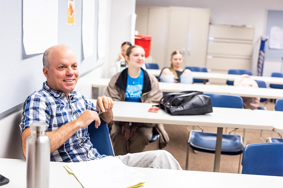 a professor at a classroom desk smiles and listens to internship presentations