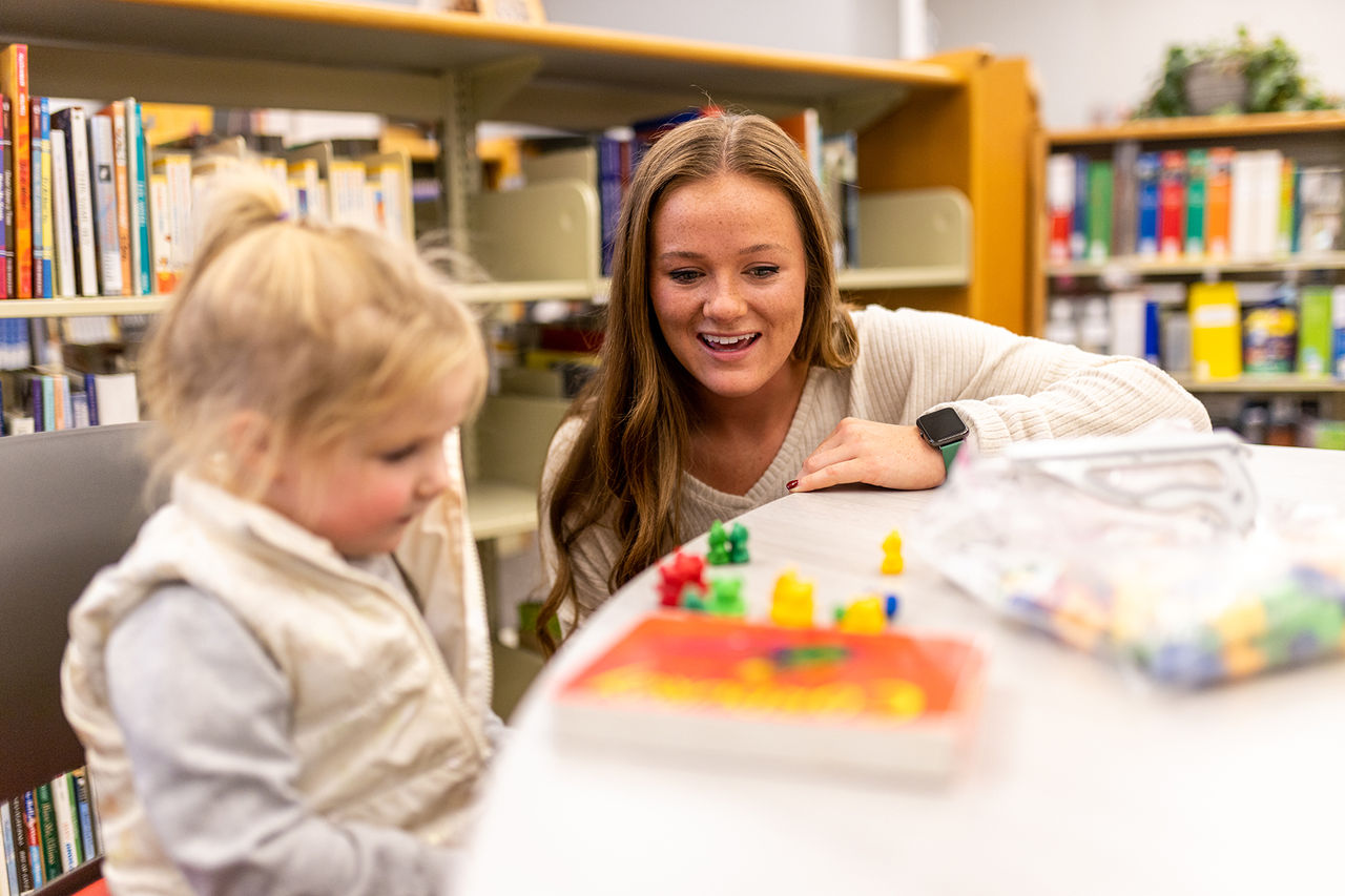 woman leaning on a table next to a small child with small colorful toys on the table in front of her