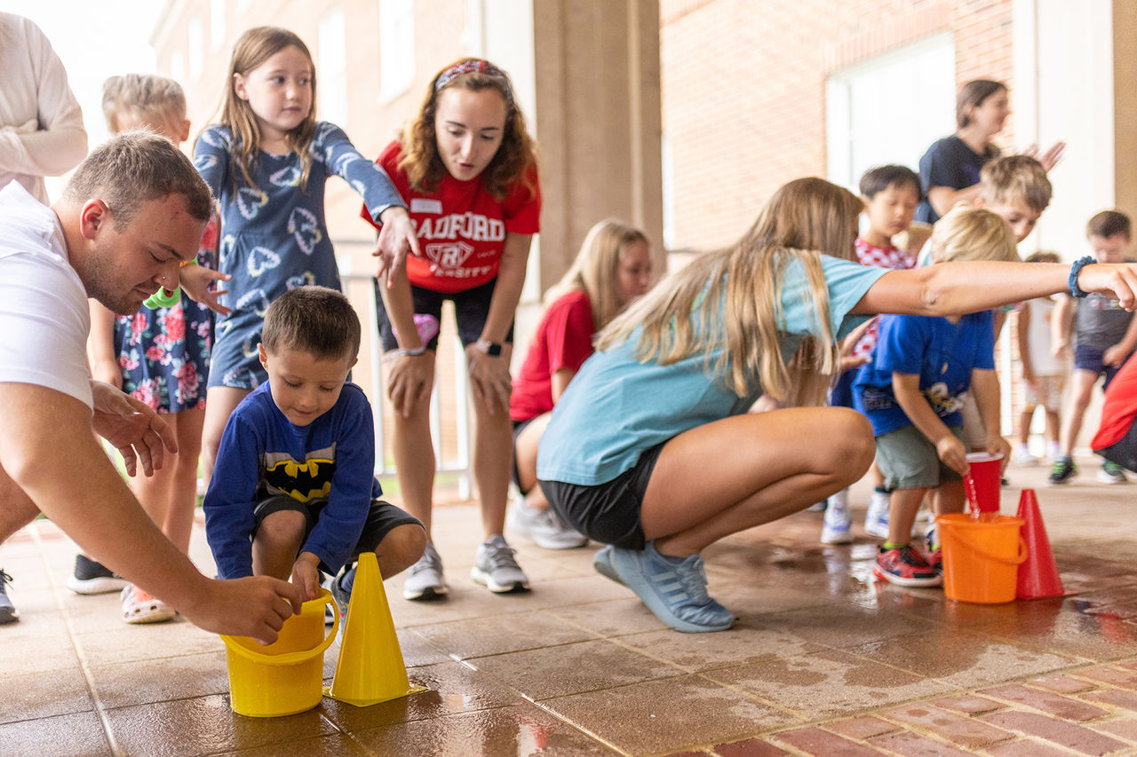 Radford students leading a children's relay race where they fill up a cup from a bucket and run with it 