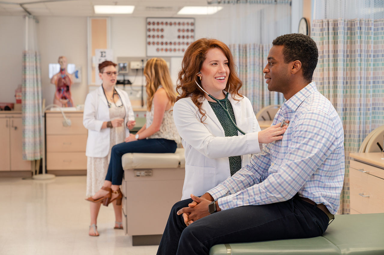 student in white lab coat and wearing a stethoscope hearing the heartbeat of a patient